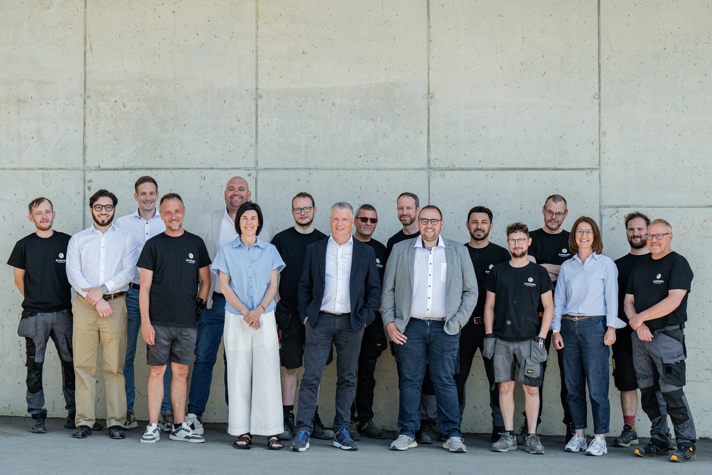 Photo de groupe des équipes Amecam Van Conversion, atelier en t-shirt noir et bureau en chemise, devant le mur de béton des ateliers de Francorchamps