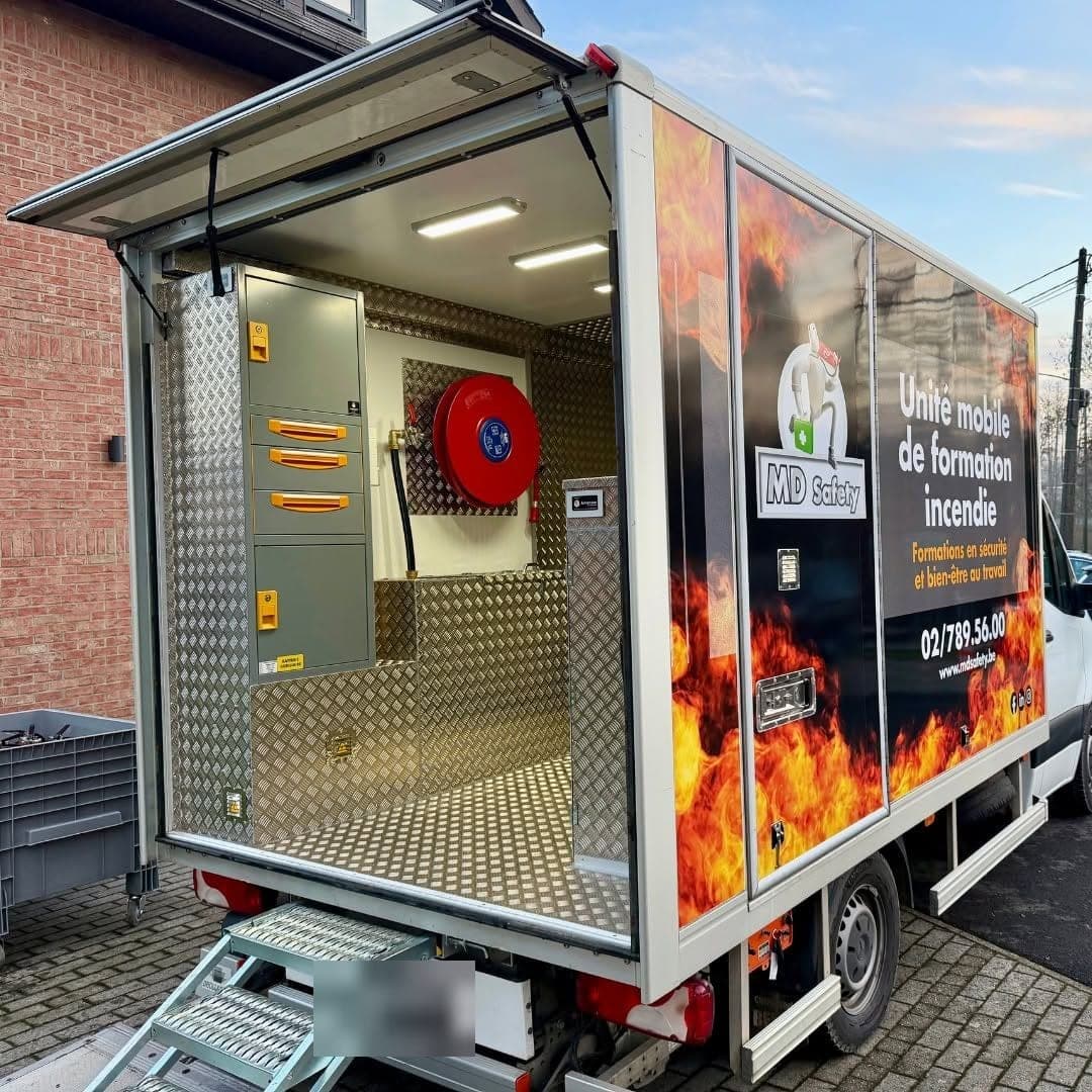 Camion MD Safety d'unité mobile de formation incendie, porte latérale relevée sur une cellule en tôle aluminium larmée, armoire grise à poignées orange et dévidoir rouge