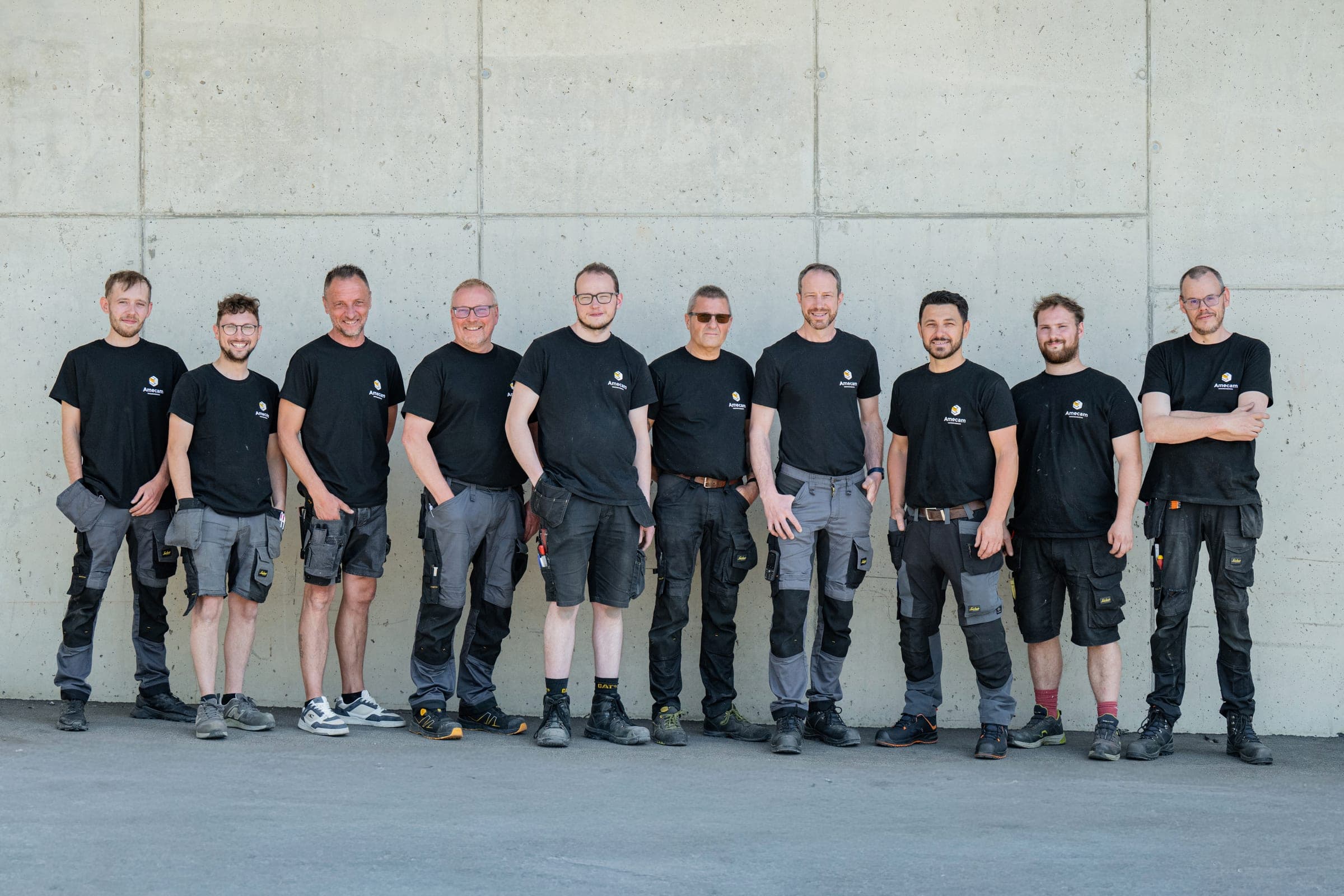 Les dix collaborateurs de l’atelier Amecam Van Conversion en t-shirt noir et pantalon de travail, devant les ateliers de Francorchamps