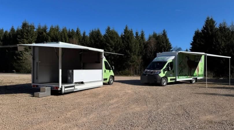 Deux camions Anim'mobil du Parc naturel Hautes Fagnes-Eifel sur un terrain en forêt, l'un ouvert sur son espace d'accueil, l'autre avec son auvent déployé