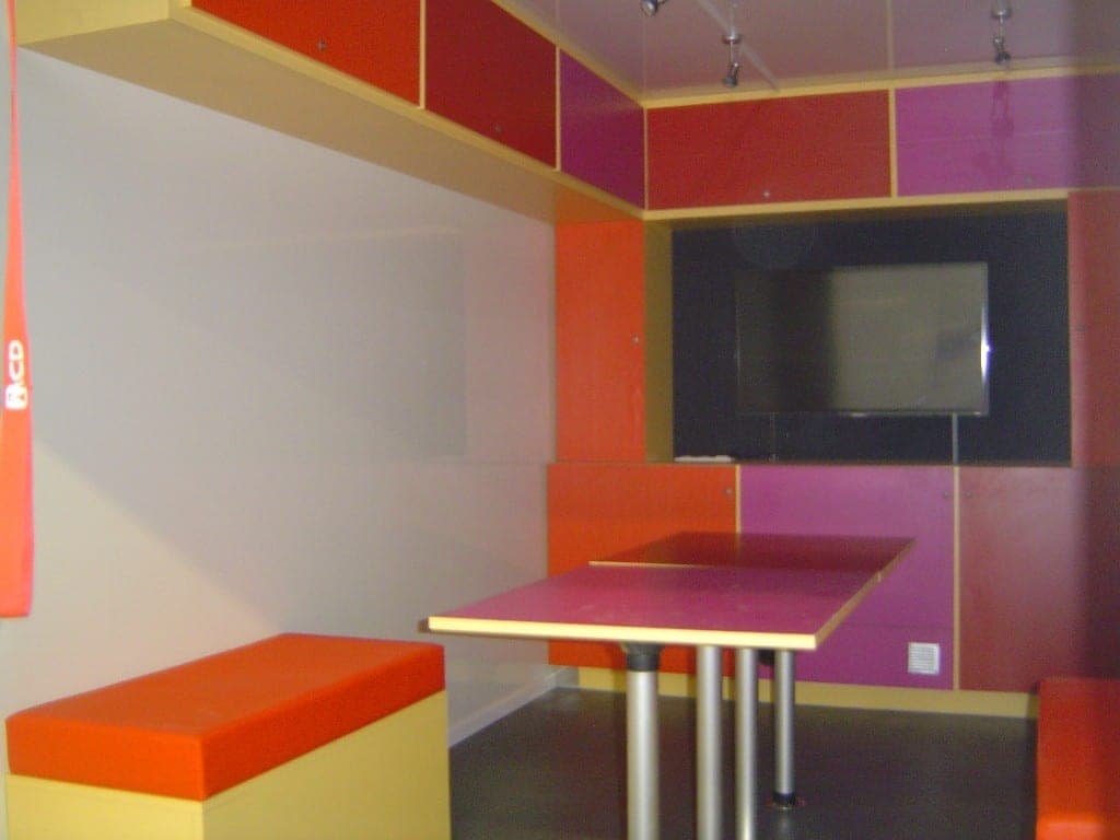 Espace de réunion coloré dans une unité mobile : banquette orange, table haute et écran mural encastré