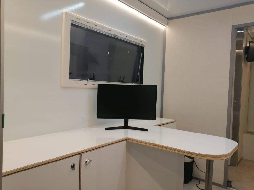 Bureau mobile avec plan de travail blanc en angle, écran sur pied et fenêtre donnant sur l’extérieur