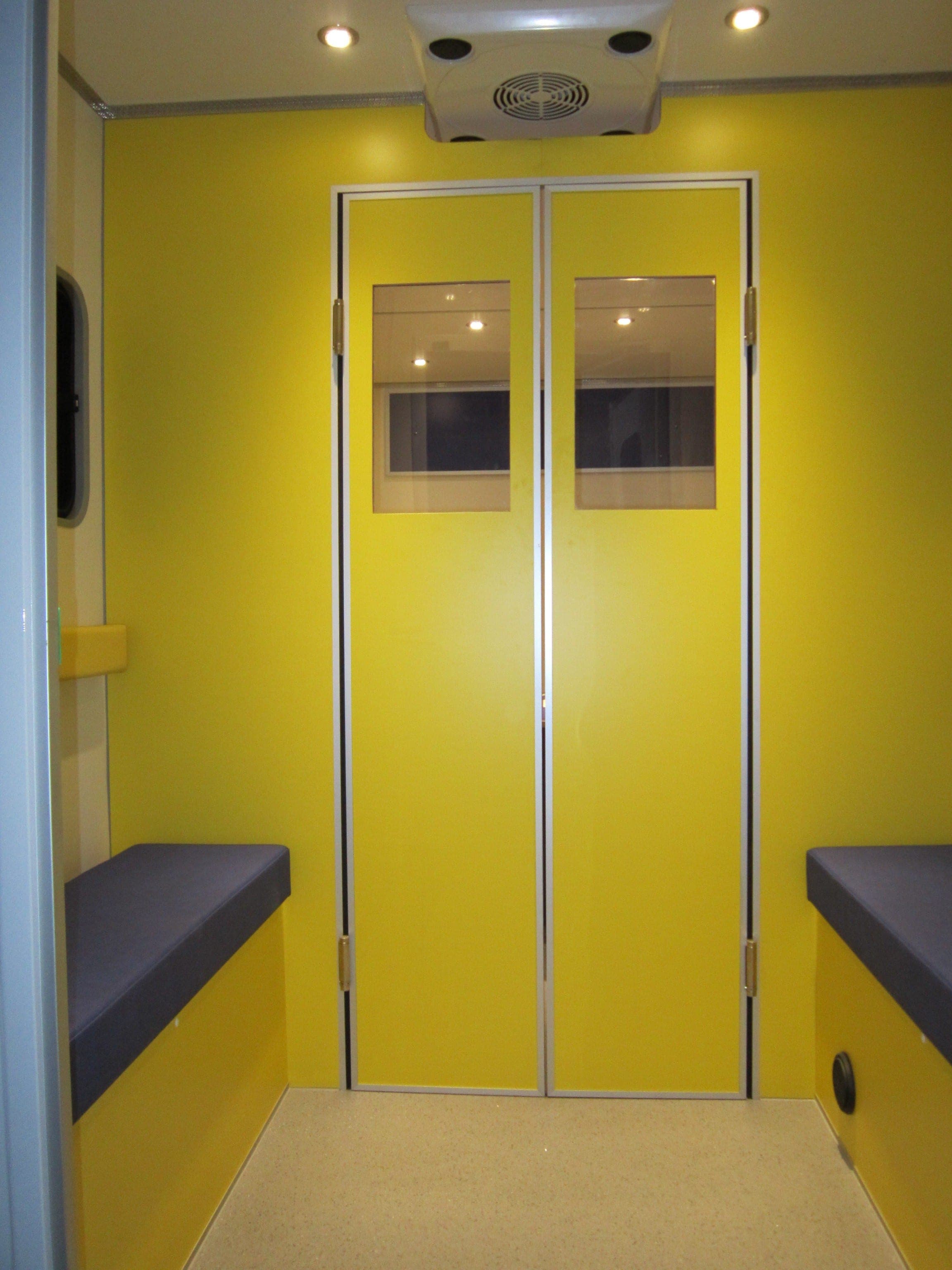 Intérieur d’une unité mobile : doubles portes vitrées jaunes, banquettes violettes et plafonnier de ventilation