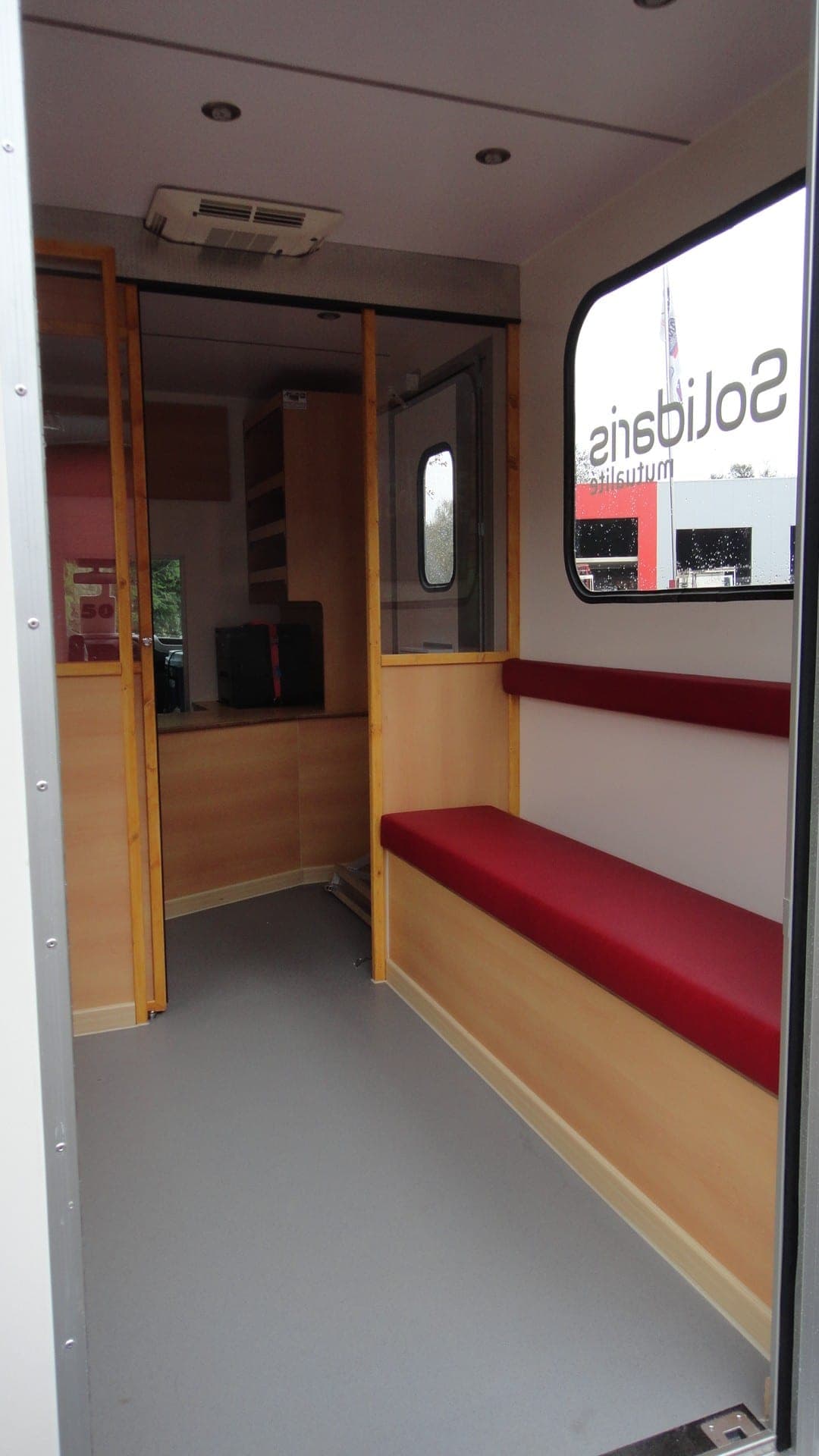 Bureau mobile d’une mutualité : sas d’entrée, banquette rouge, cloison en bois clair et guichet vitré