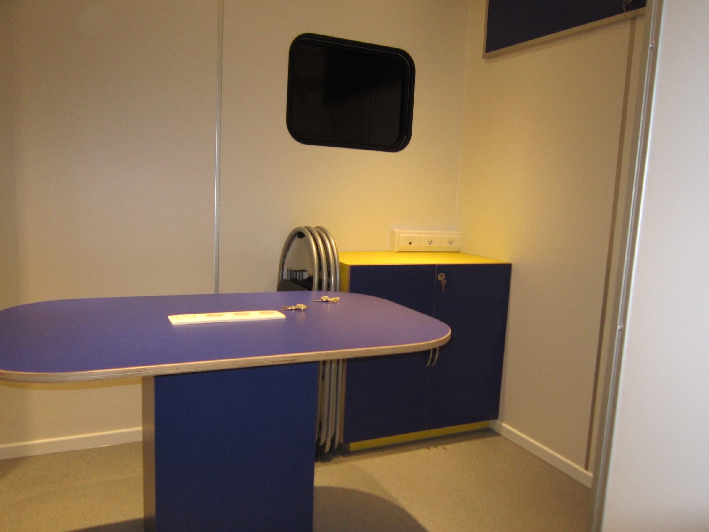Salle de réunion d’une unité mobile : table ovale violette, chaises pliantes rangées et écran mural
