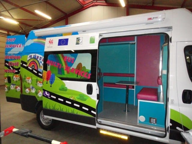 Bus multi-services habillé d’un décor illustré, porte latérale ouverte sur la banquette et la table de l’espace d’accueil