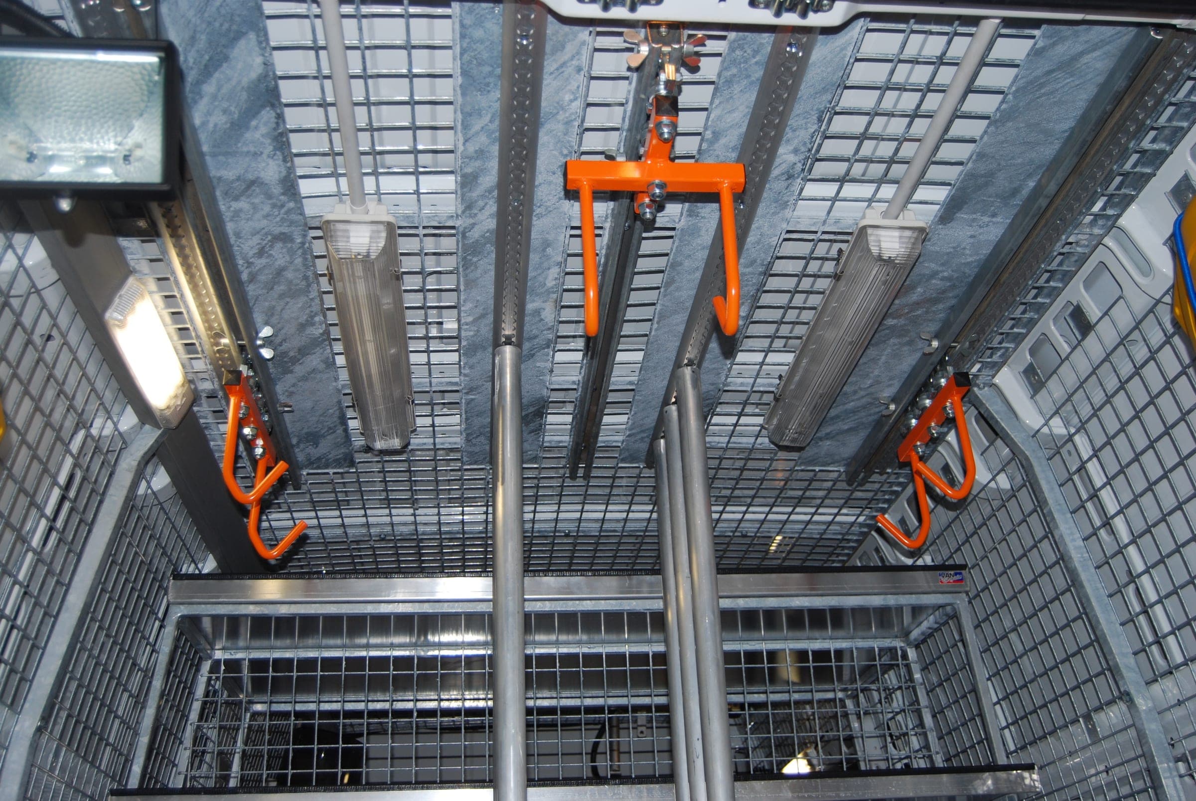 Vue plongeante dans un fourgon d’assistance grillagé : palan orange, tubes de rangement et projecteur de travail