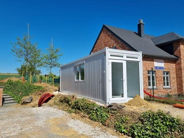 Module de bureau préfabriqué gris posé sur un chantier, porte vitrée et fenêtre, à côté d’une maison en briques