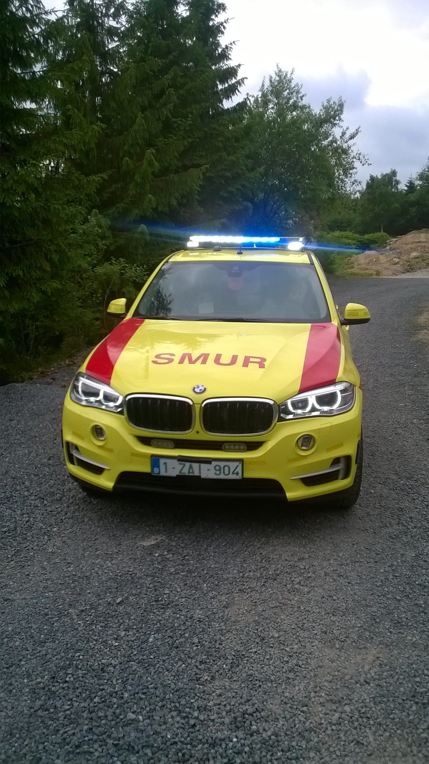 Véhicule SMUR jaune avec rampe lumineuse bleue en toiture, vu de face sur un chemin bordé de sapins