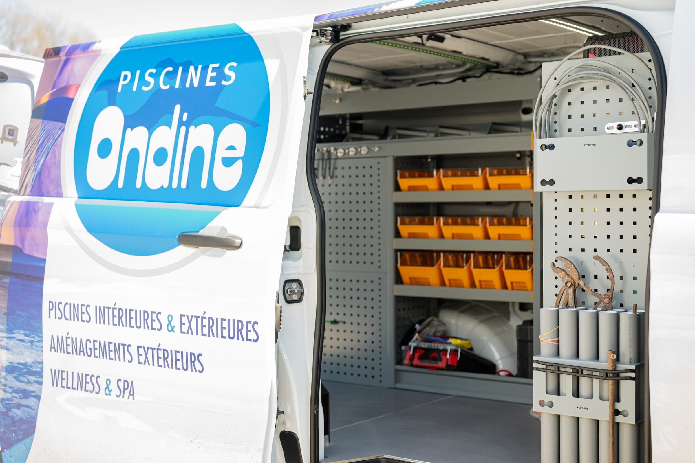 Fourgon lettré d’un installateur de piscines, porte latérale ouverte sur un rayonnage à bacs orange et des tubes de rangement