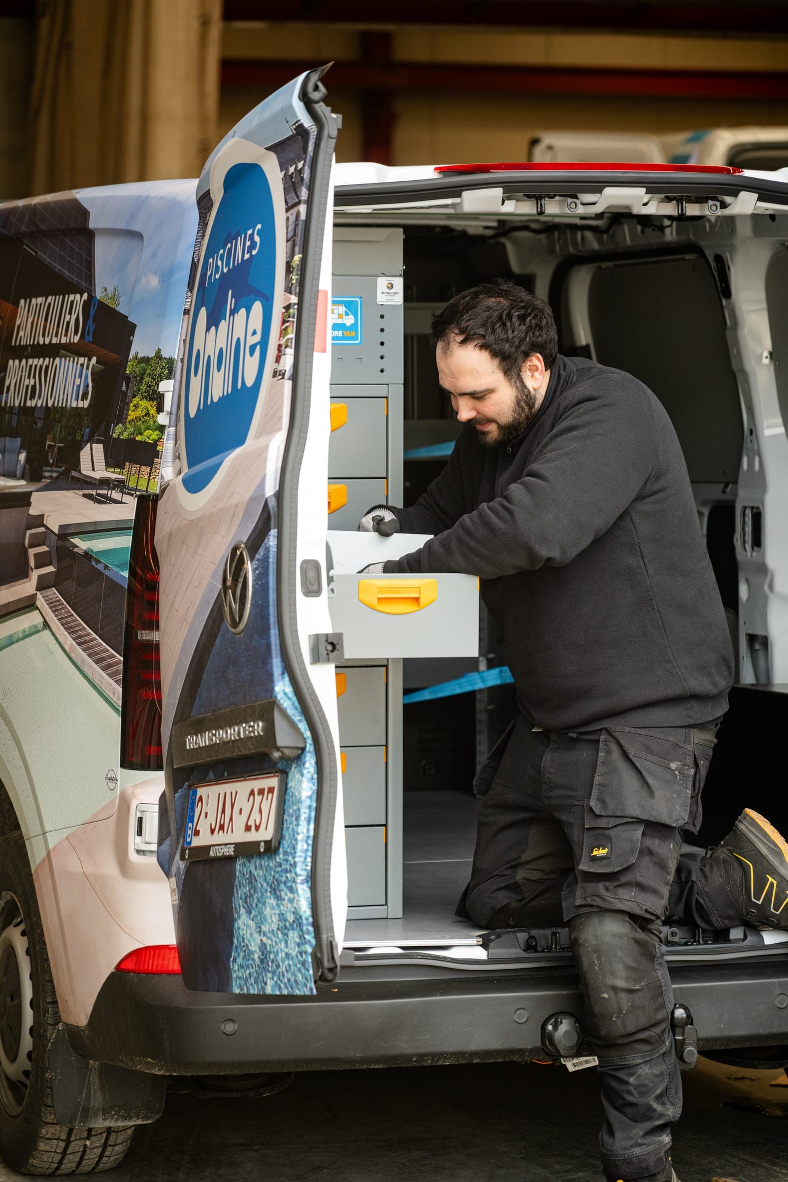 Technicien Amecam agenouillé dans un Volkswagen Transporter aux couleurs de Piscines Ondine, tiroir de rangement ouvert